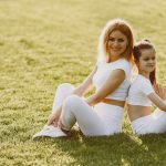 A mother and daughter sit back to back in matching activewear on a sunny grass field, enjoying a summer day together.