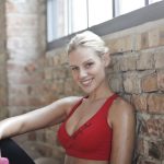 Blonde woman in red sportswear smiling with a dumbbell in a bright gym setting.