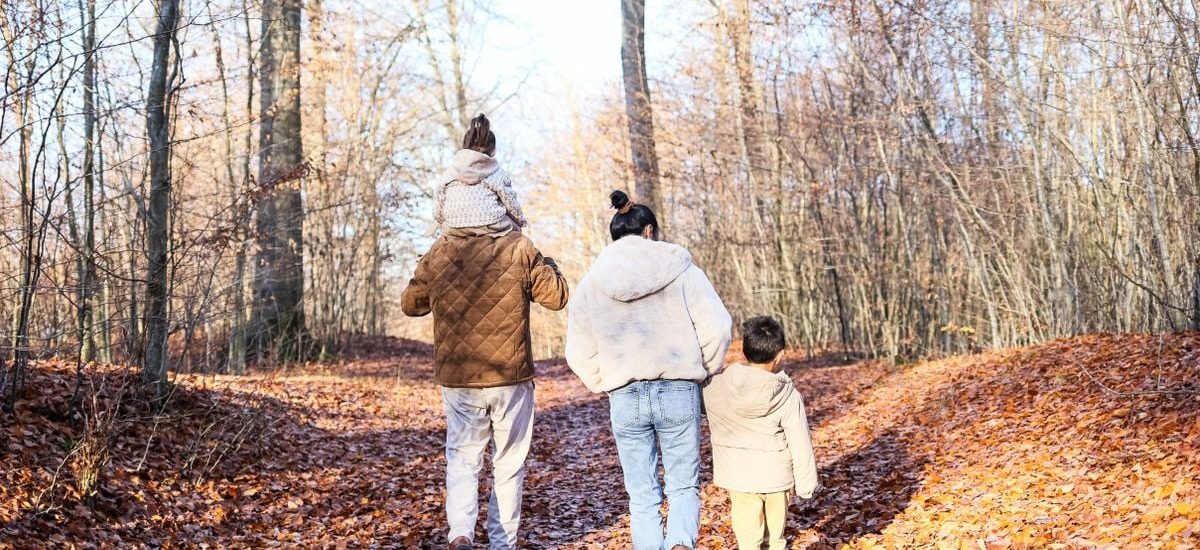 A family of four strolling on a leaf-covered path in a fall forest.
