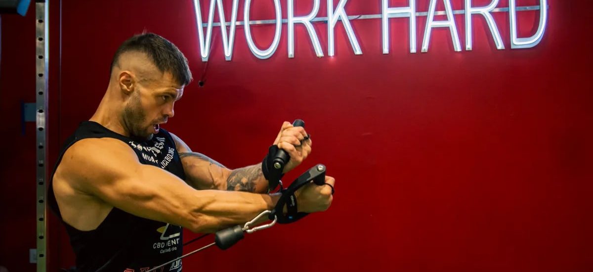 A muscular man exercising in the gym with a motivational 'Work Hard' neon sign on a red background.